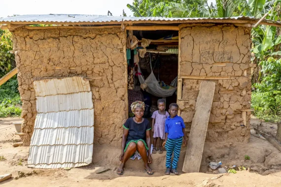 Kvinna med två barn framför familjens enkla hus i Ghana.